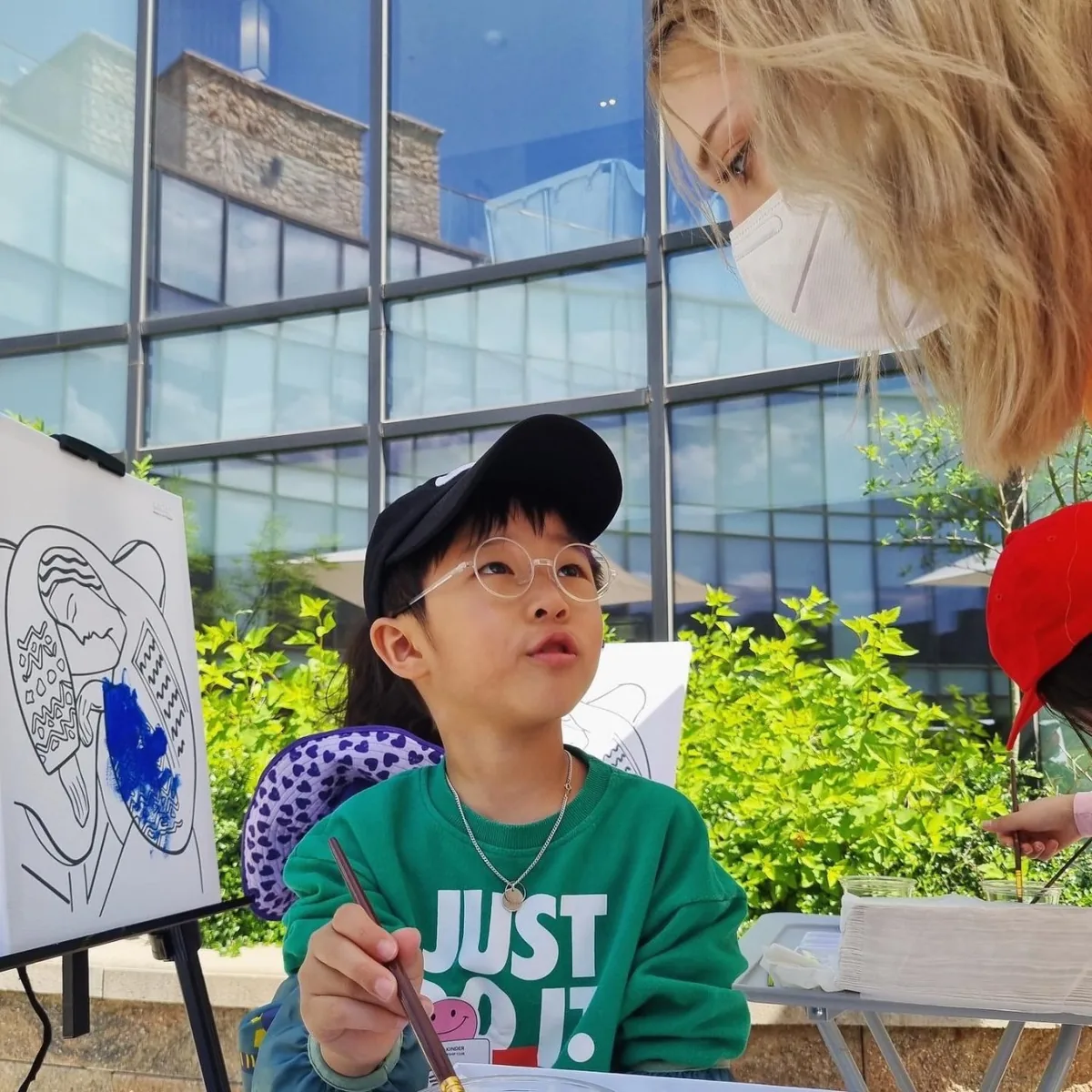 야외 이젤 앞에서 붓을 들고 선생님을 올려다보는 아이